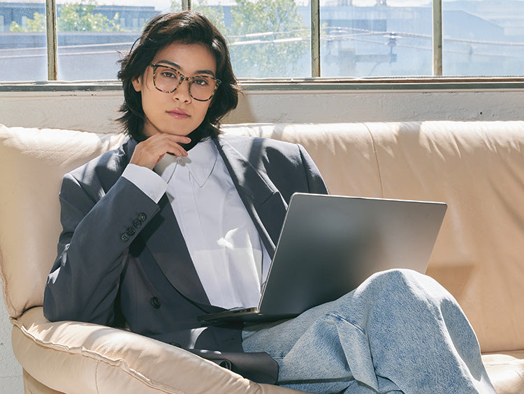 Person in glasses sitting on a couch with a laptop, wearing a casual blazer indoors.