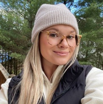 A person wearing glasses and a beanie, smiling outdoors with trees in the background.