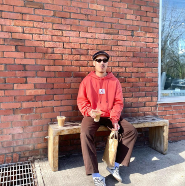 Person sitting on a bench with iced coffee, wearing a red hoodie and sunglasses, against a brick wall.